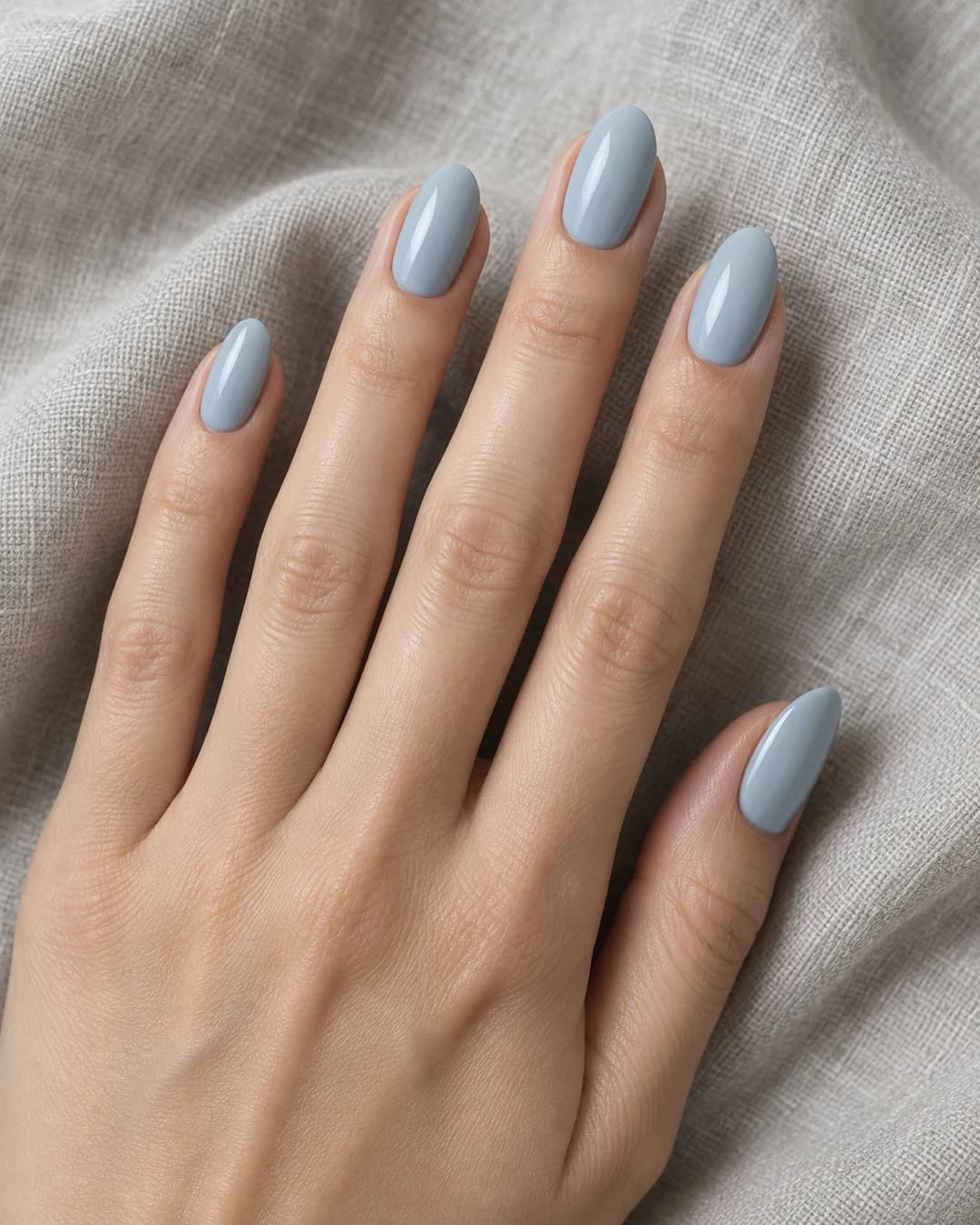 Mano de piel clara con uñas almendradas medias en esmalte cremoso azul niebla de subtono gris.