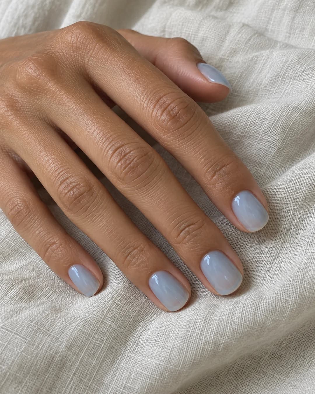 Mano de piel tostada media con uñas ovaladas cortas en una capa translúcida azul gris que deja ver la línea natural.