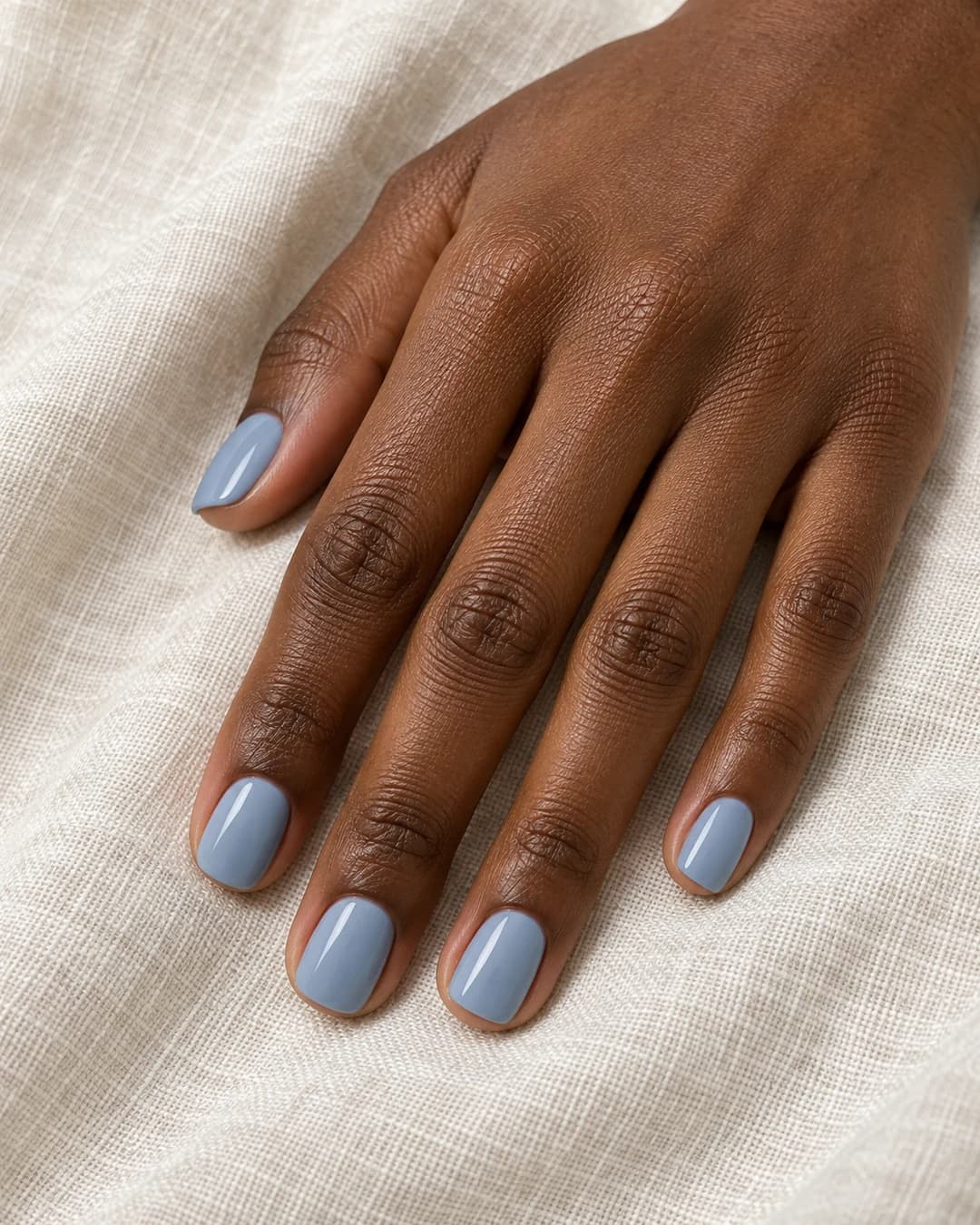 Mano de piel marrón profunda con uñas squoval cortas en esmalte azul grisáceo suave y brillante.