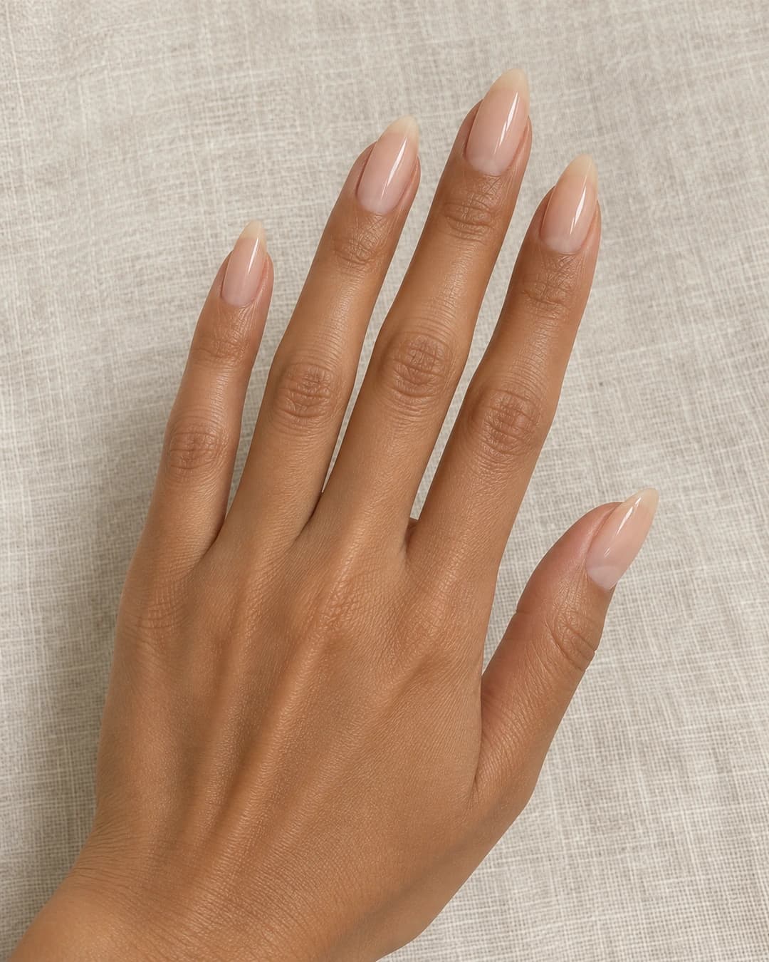 Mano adulta de piel marrón clara con uñas almendradas largas y esmalte nude cálido translúcido.