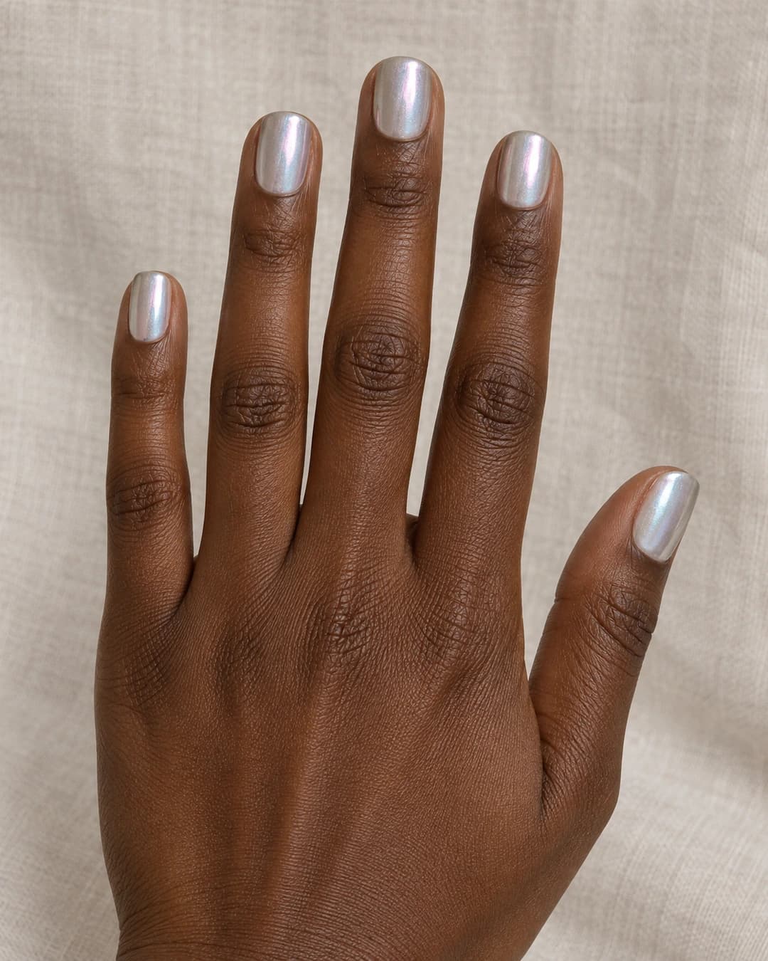 Mano adulta de piel marrón profunda con uñas squoval cortas de plata nacarada e iridiscencia suave.