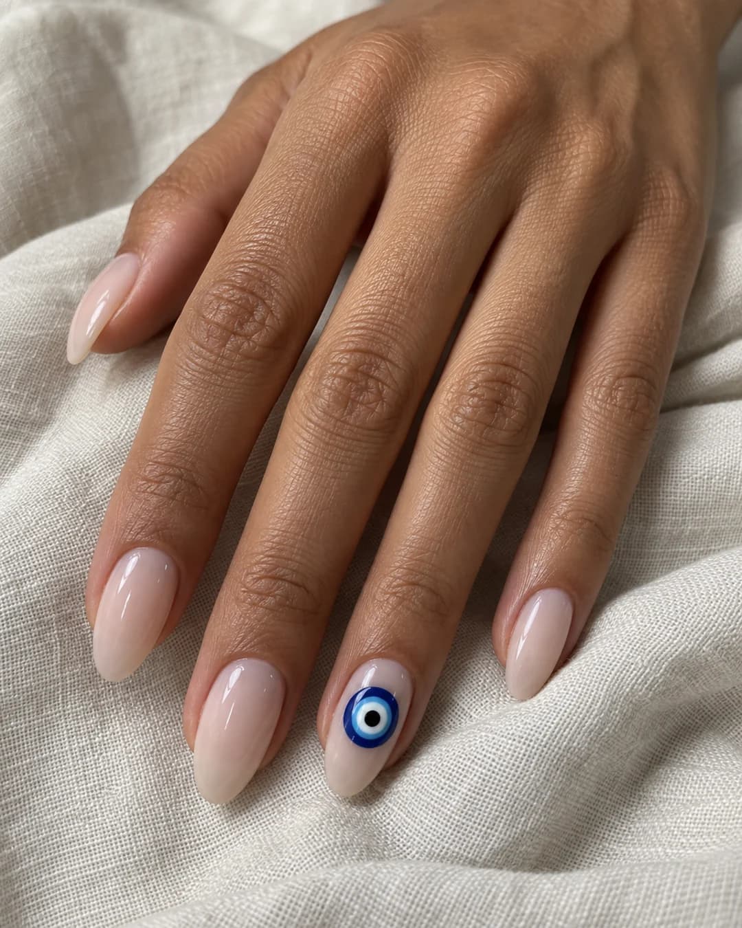 Mano adulta de piel oliva media con uñas almendradas medias en nude lechoso y un ojo turco azul en el anular.