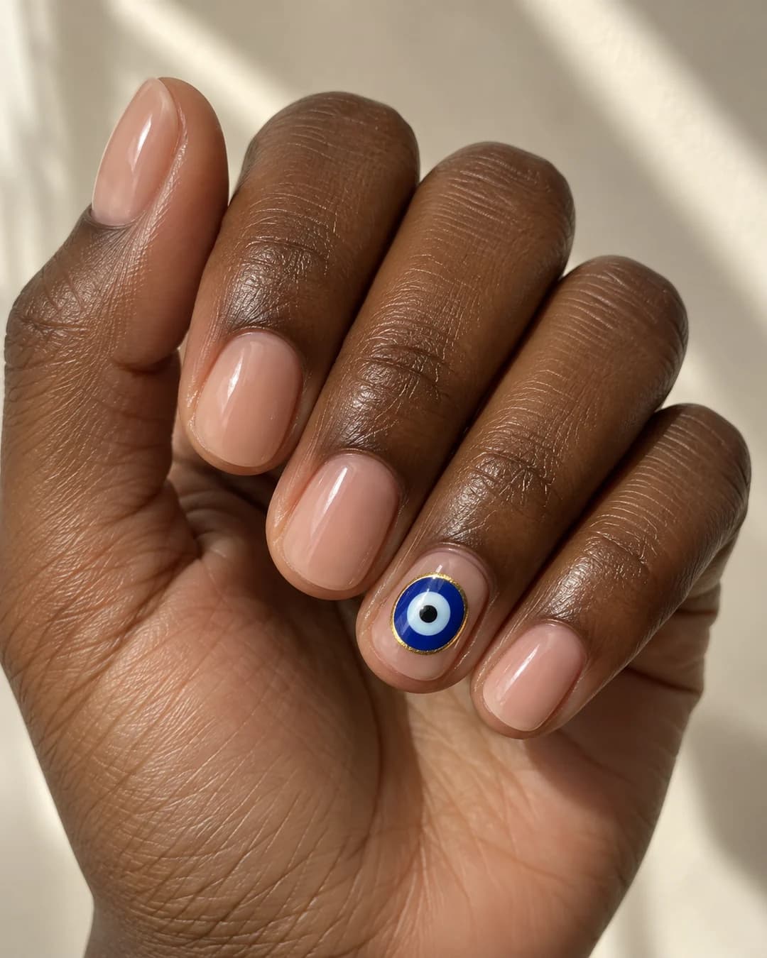 Mano adulta de piel marrón oscura con uñas cortas redondas nude y un ojo turco azul con filo dorado en el anular.