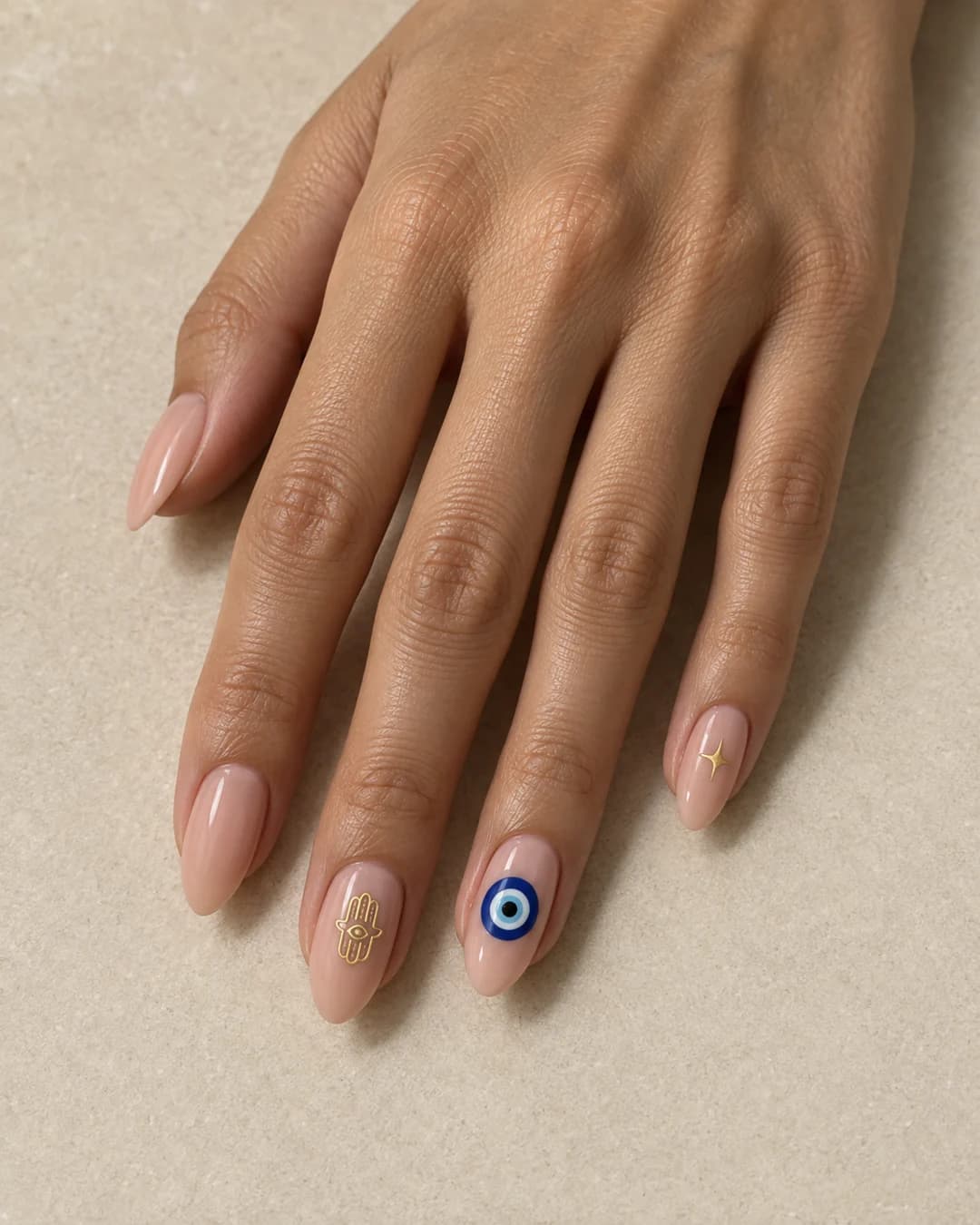 Mano adulta de piel oliva con uñas almendradas medias nude y tres detalles: ojo turco azul, hamsa dorada y estrella fina.