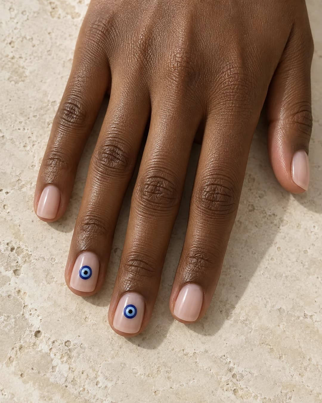 Mano adulta de piel marrón profunda con uñas cortas squoval nude lechoso y dos ojos turcos azules pequeños en los dedos medio y anular.