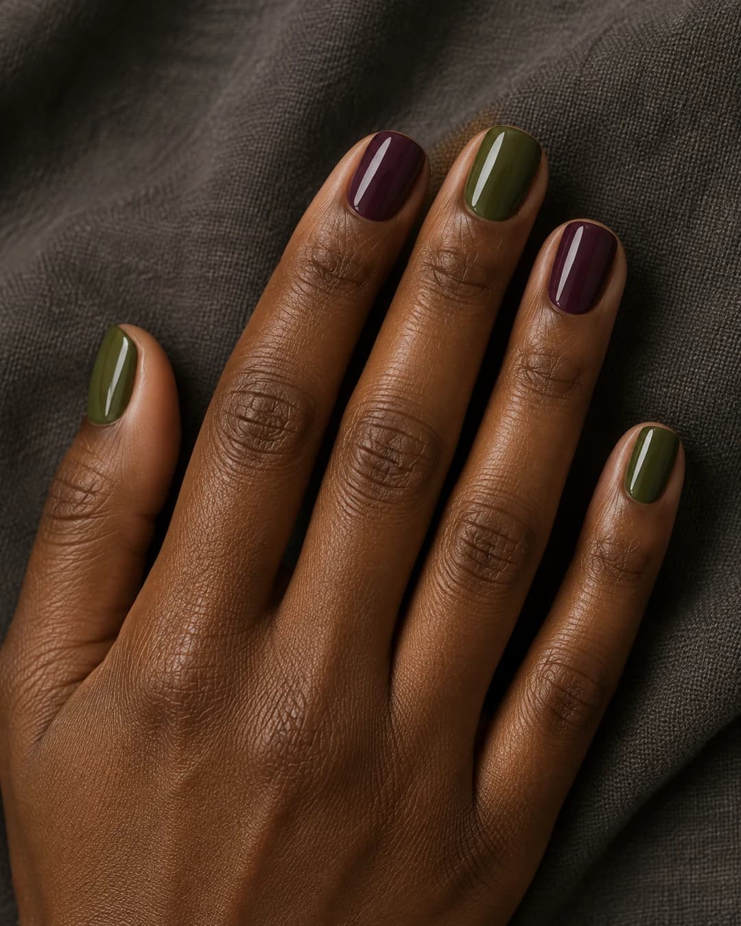 Uñas squoval cortas alternadas en verde oliva y ciruela sobre piel profunda.
