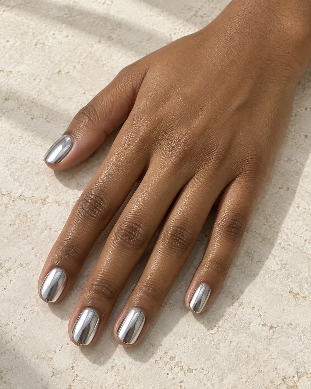 Mano adulta de piel oliva con cinco uñas ovaladas cortas en plata fría muy reflectante.