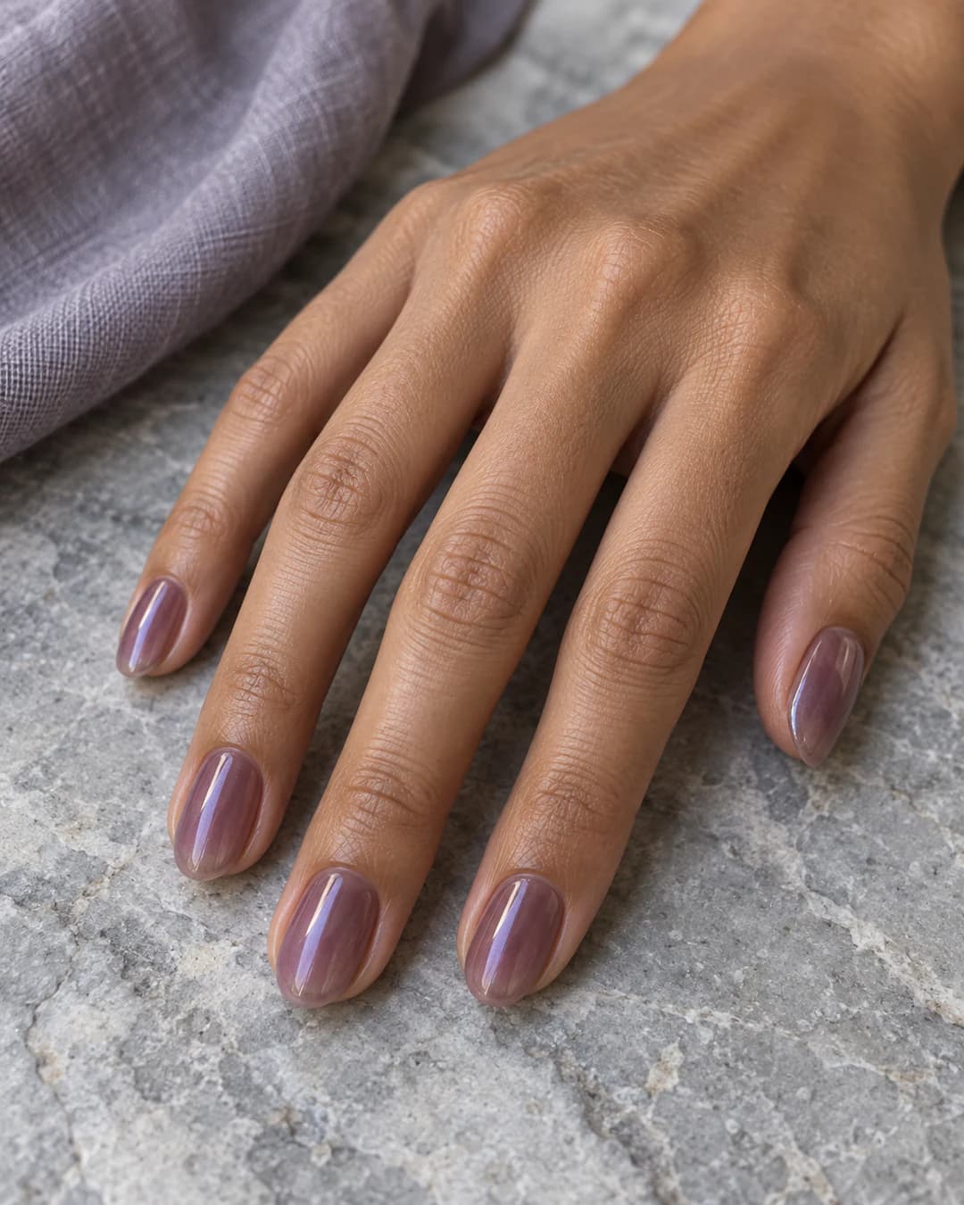 Mano de piel marrón media con uñas almendradas cortas en malva translúcido con reflejo violeta y brillo alto.