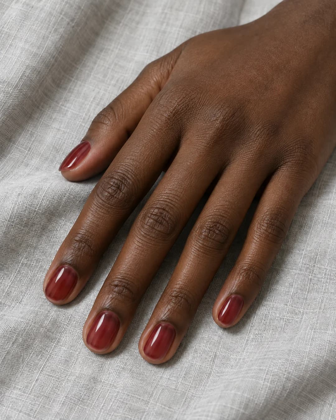 Mano de piel oscura con uñas cortas ovaladas en cereza translúcida y acabado brillante.