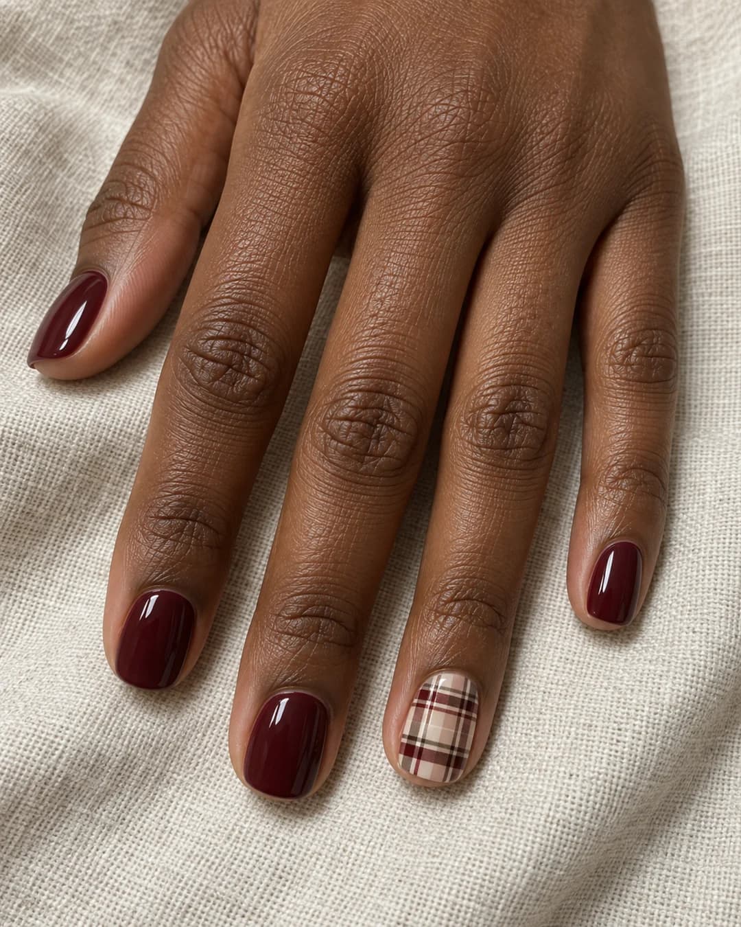 Mano de piel marrón oscura con uñas squoval cortas color vino y un acento tartán marfil, cacao y merlot.