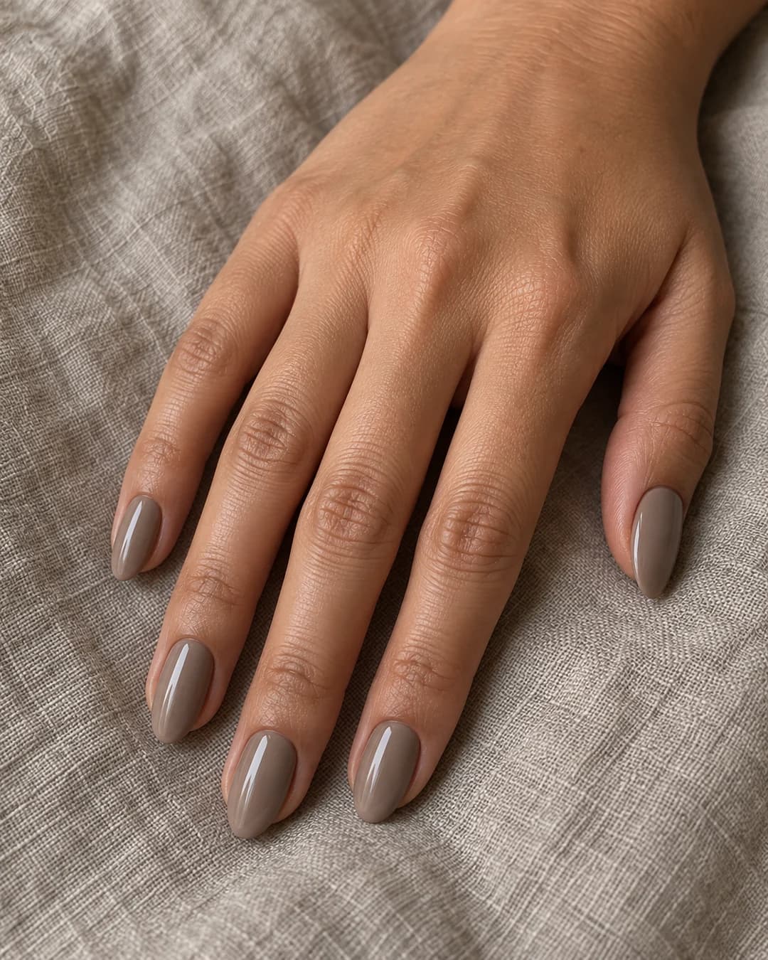 Mano de piel media con cinco uñas almendradas medias en esmalte taupe cremoso marrón-gris brillante.