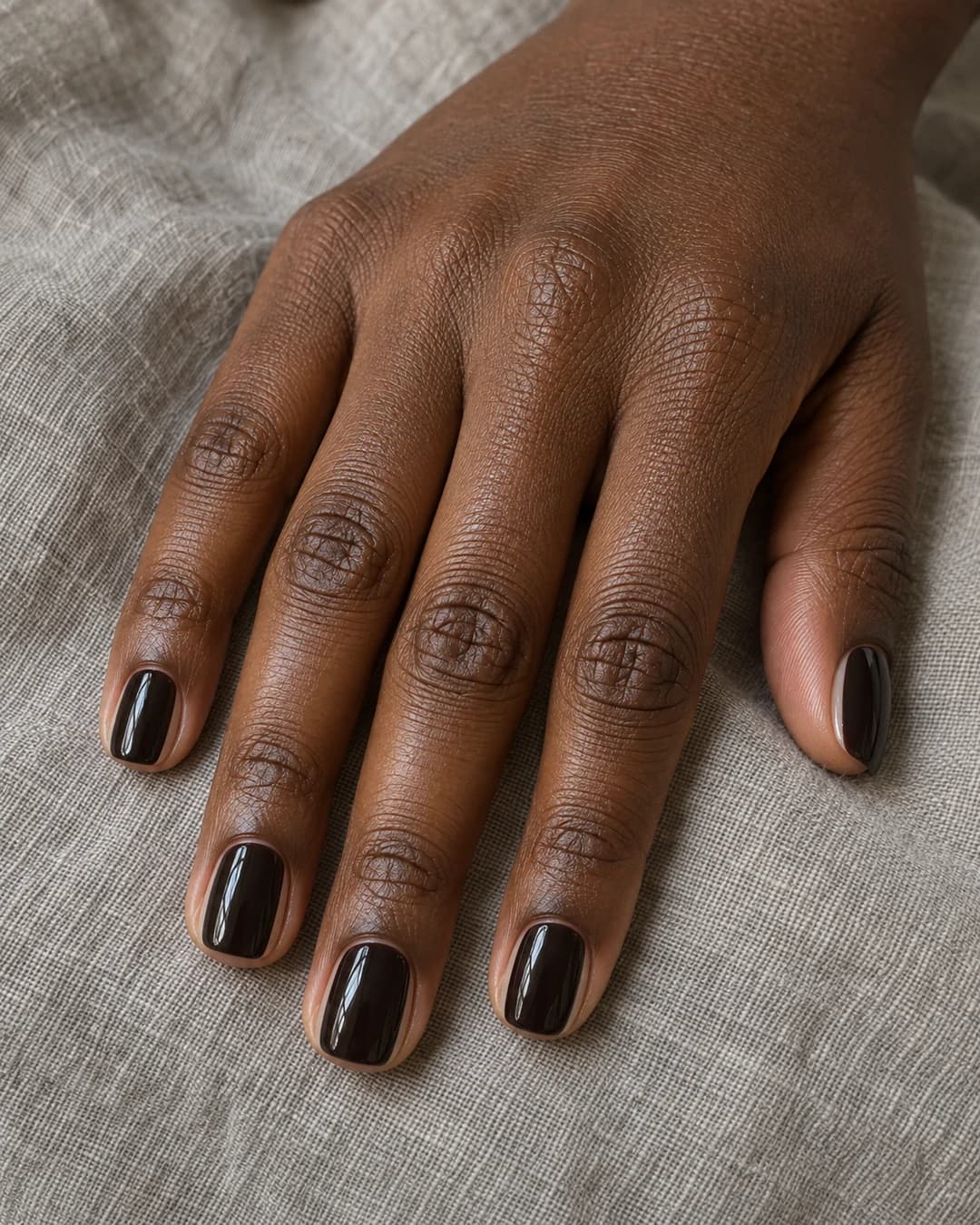 Mano adulta de piel marrón profunda con cinco uñas squoval cortas en espresso casi negro y márgenes laterales naturales visibles.