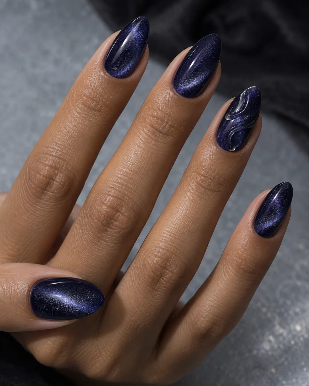 Mano adulta de piel oliva con cinco uñas almendradas medias en velvet índigo magnético y dos ondas de gel transparente en una uña.