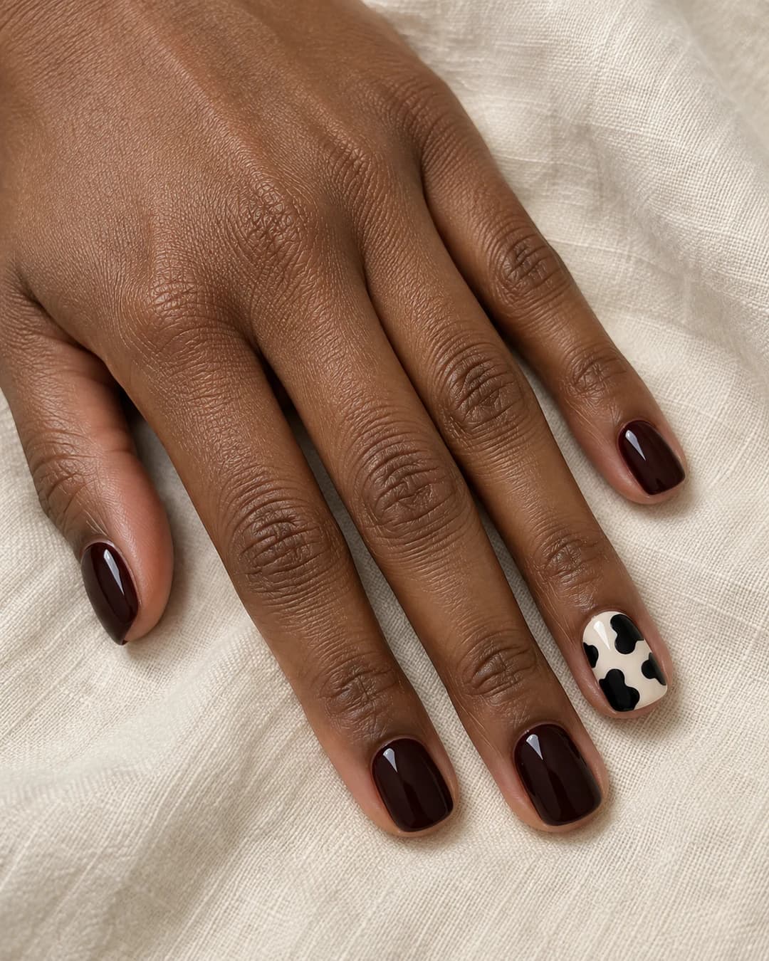 Mano de piel marrón oscura con uñas squoval cortas black cherry y un acento marfil con manchas negras grandes de vaca.