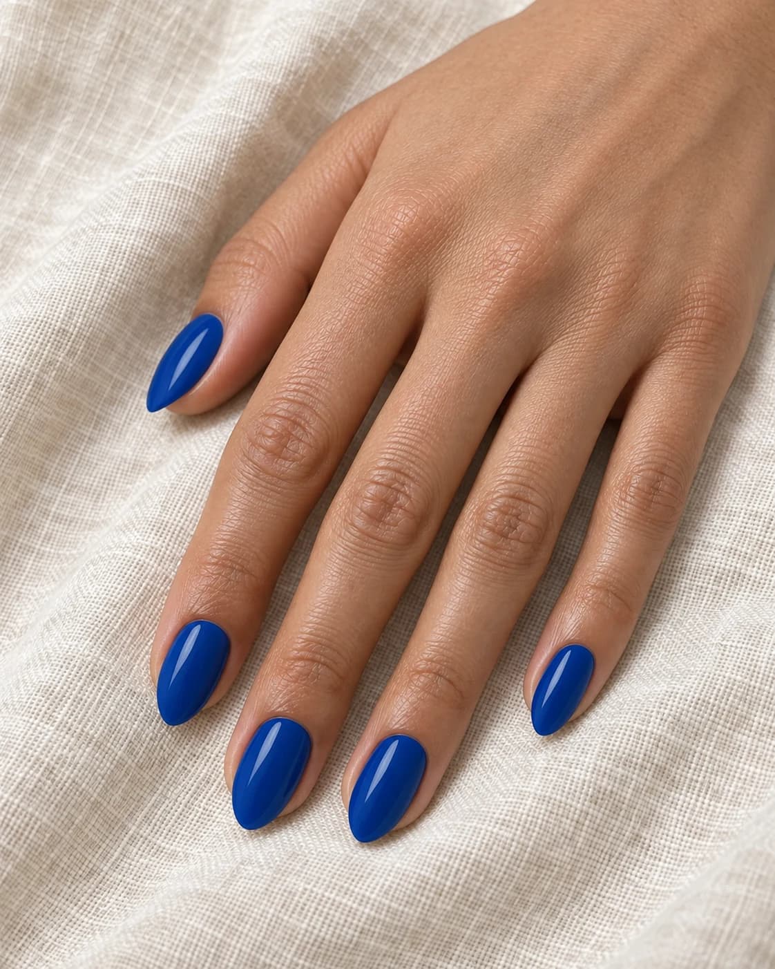 Mano de piel clara con cinco uñas almendradas medias en esmalte azul zafiro intenso y brillante.