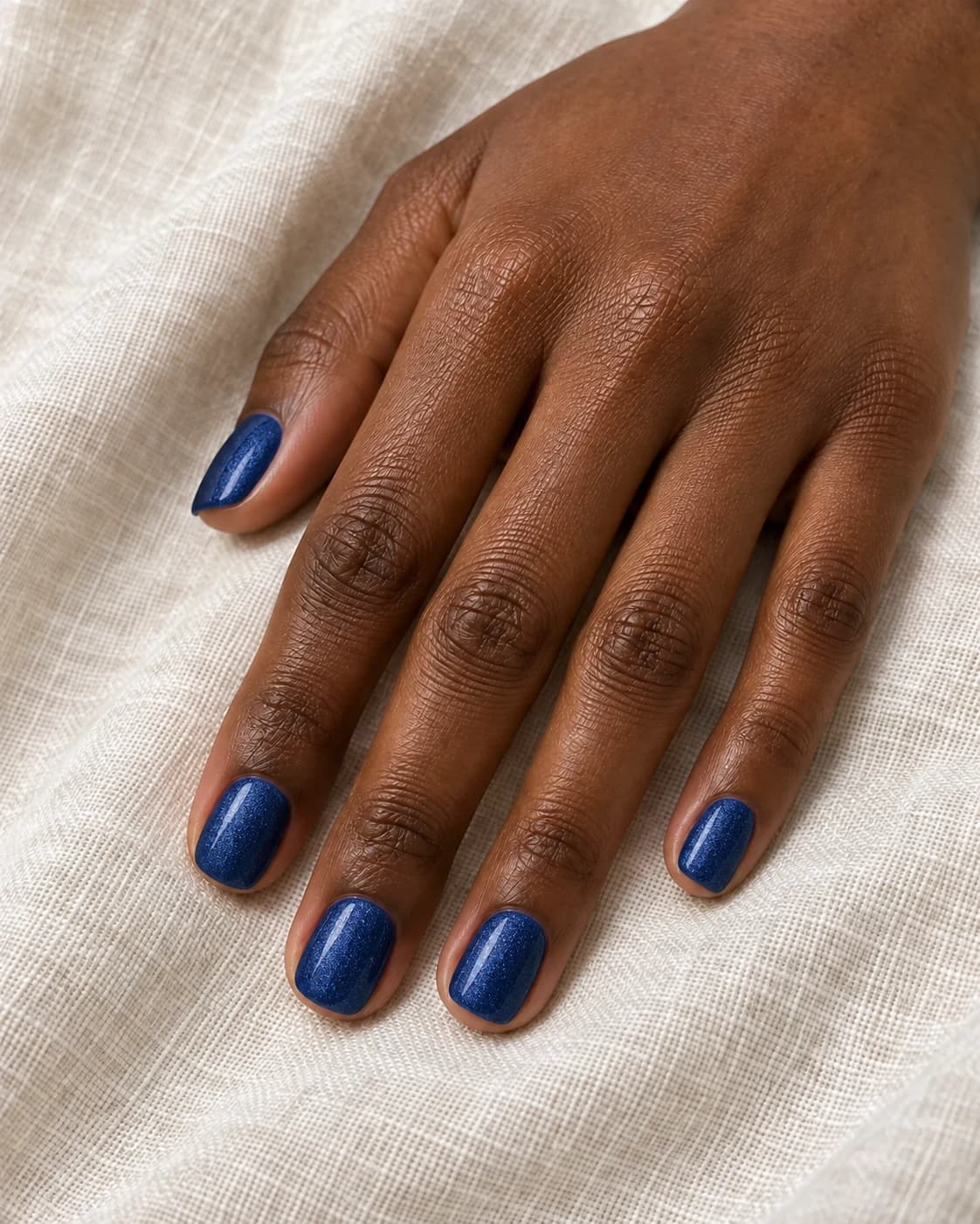 Mano de piel morena profunda con cinco uñas squoval cortas en azul zafiro con microbrillo fino.