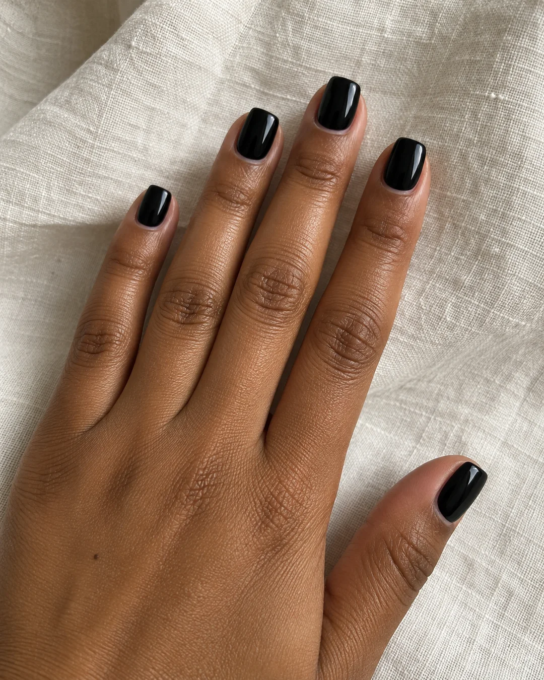 Mano adulta de piel marrón con cinco uñas negras brillantes cortas, laterales ligeramente afinados y puntas planas sobre lino crema.
