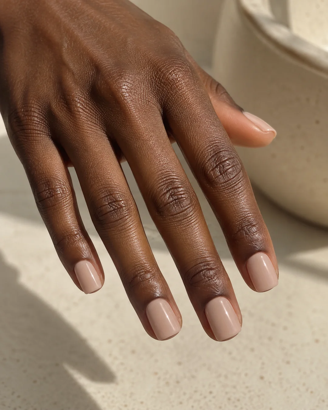 Mano adulta de piel oscura con cinco uñas nude beige brillantes, cortas y cuadradas sobre cerámica crema.