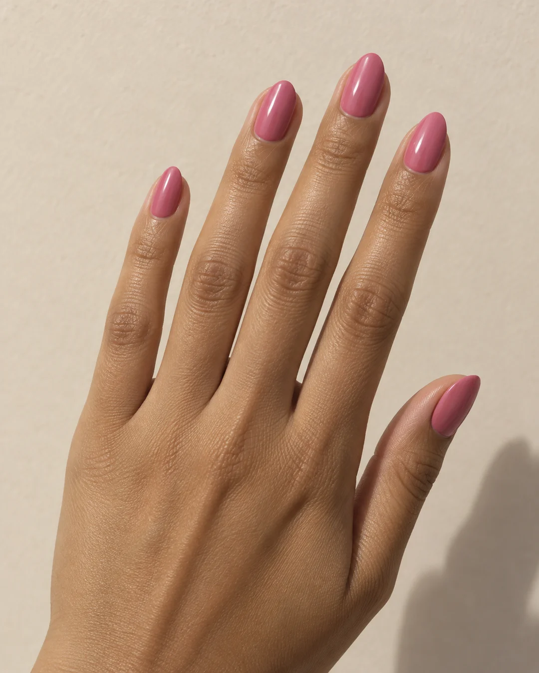 Mano adulta de piel oliva con cinco uñas cortas almendradas en rosa liso brillante sobre papel beige claro.