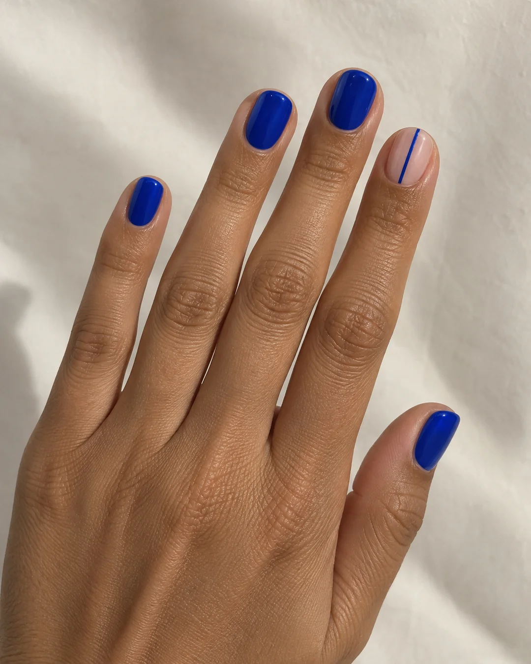 Mano adulta de piel oliva con cuatro uñas cortas azul eléctrico y una uña nude en el índice con una línea azul fina sobre papel crema.