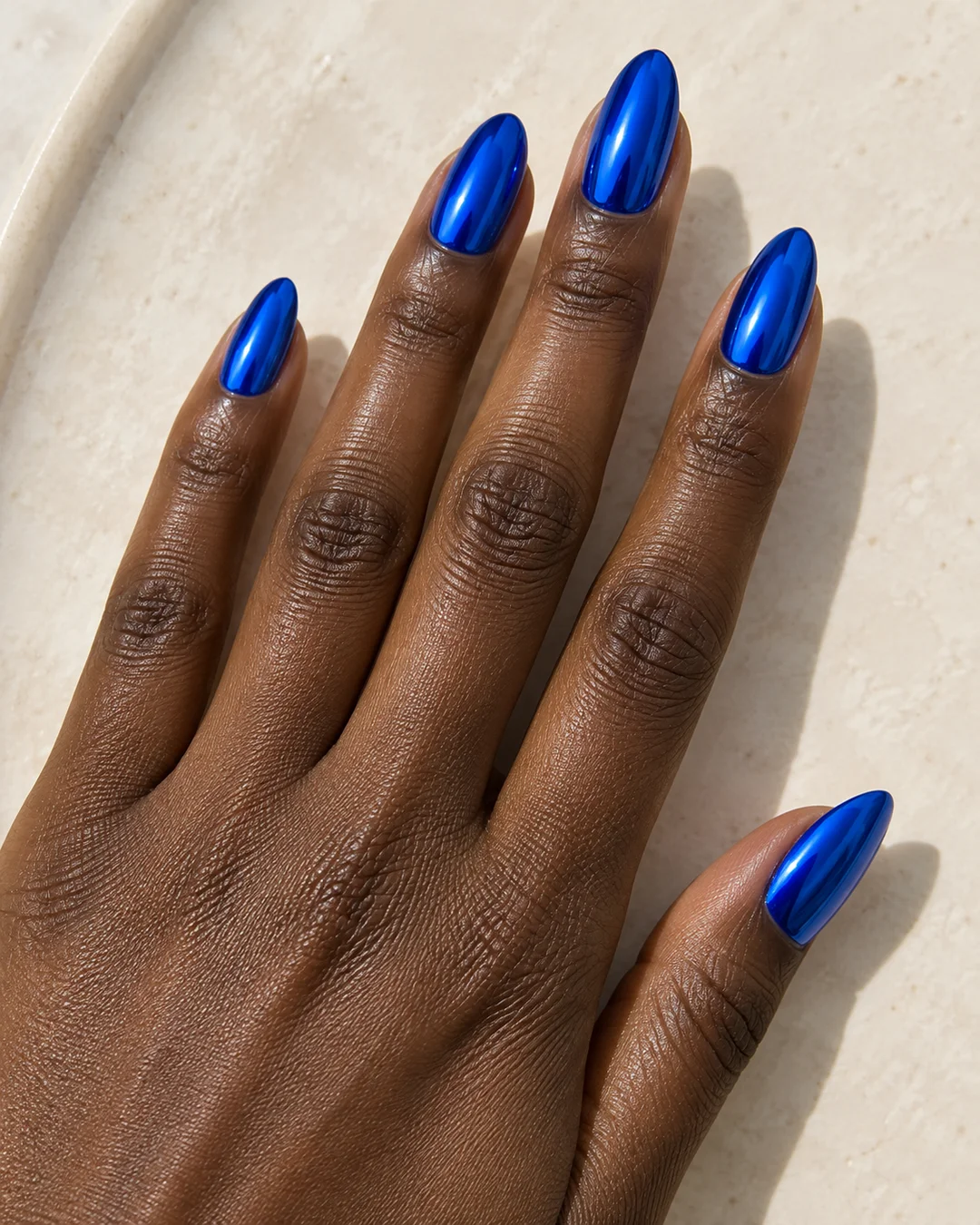 Mano adulta de piel oscura con cinco uñas almendradas medias en azul eléctrico de reflejo metálico sobre cerámica marfil.