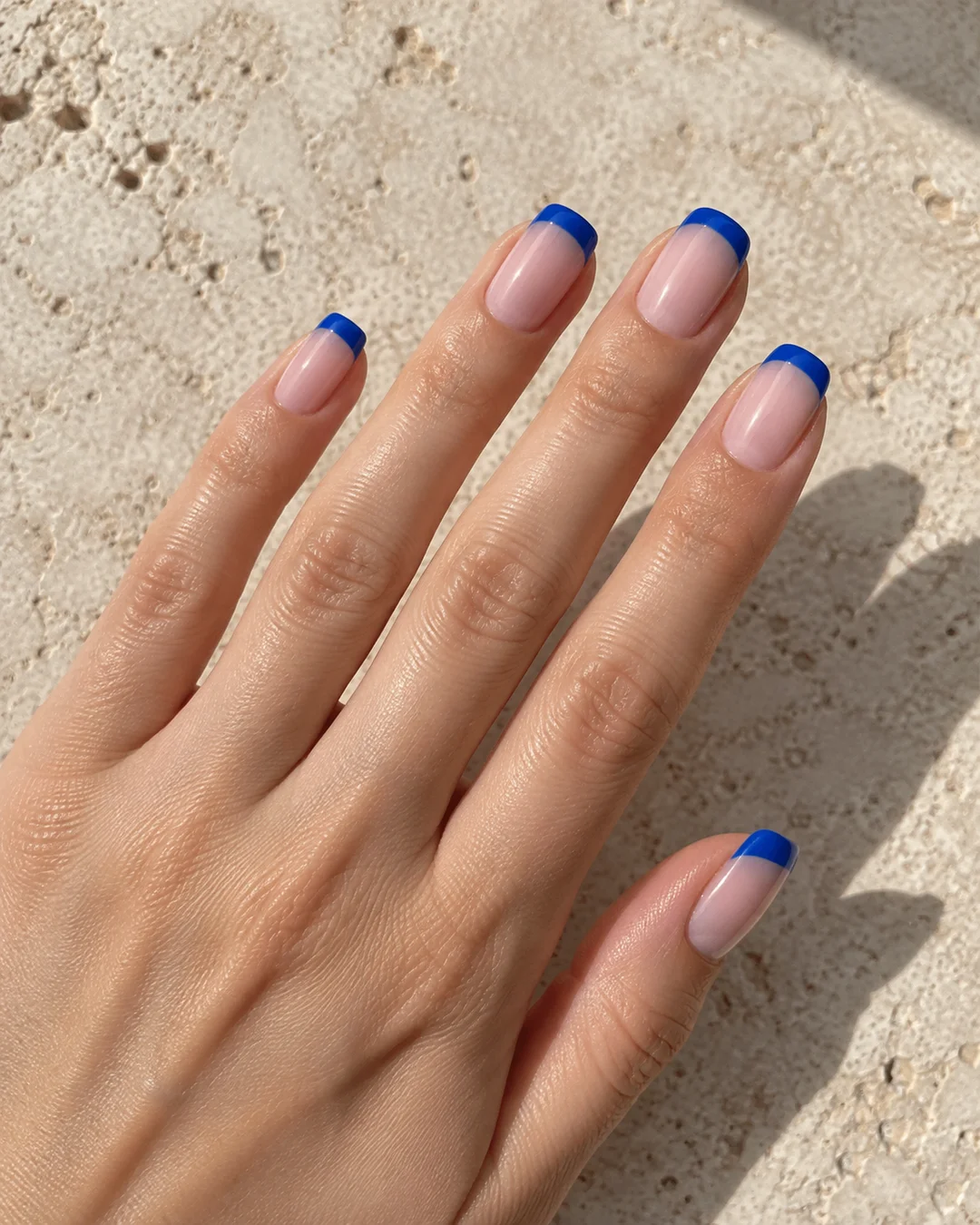 Mano adulta de piel clara con cinco uñas cuadradas medias, base nude rosada y puntas francesas azul cobalto sobre piedra beige.