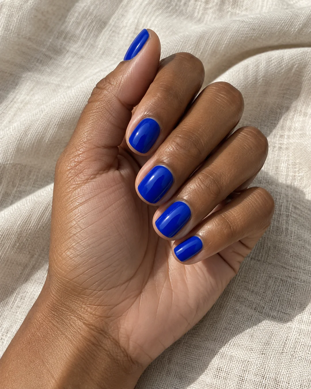 Mano adulta de piel marrón con cinco uñas squoval cortas en azul cobalto liso brillante sobre lino crema.