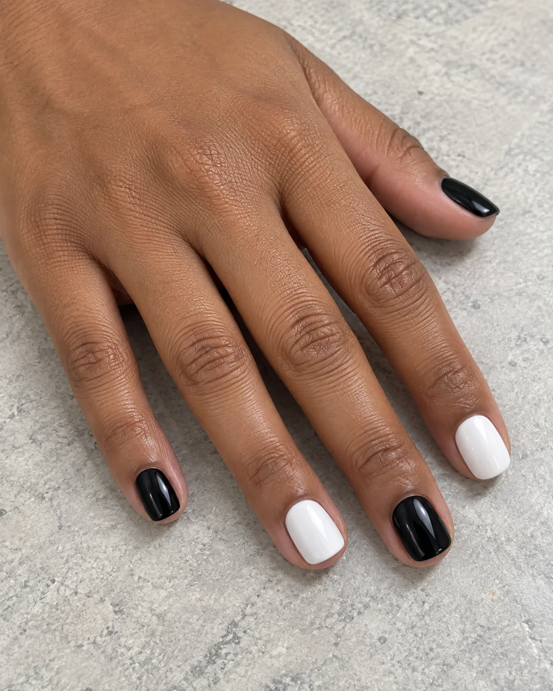 Mano de piel marrón media con uñas cortas que alternan blanco y negro lisos y brillantes.