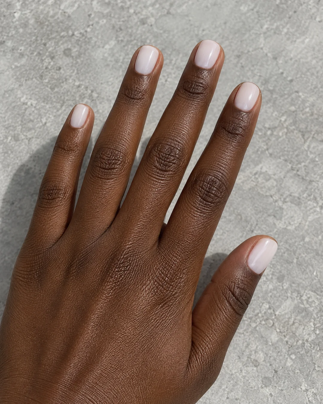 Mano adulta de piel oscura con cinco uñas squoval muy cortas en blanco lechoso brillante sobre piedra gris clara.