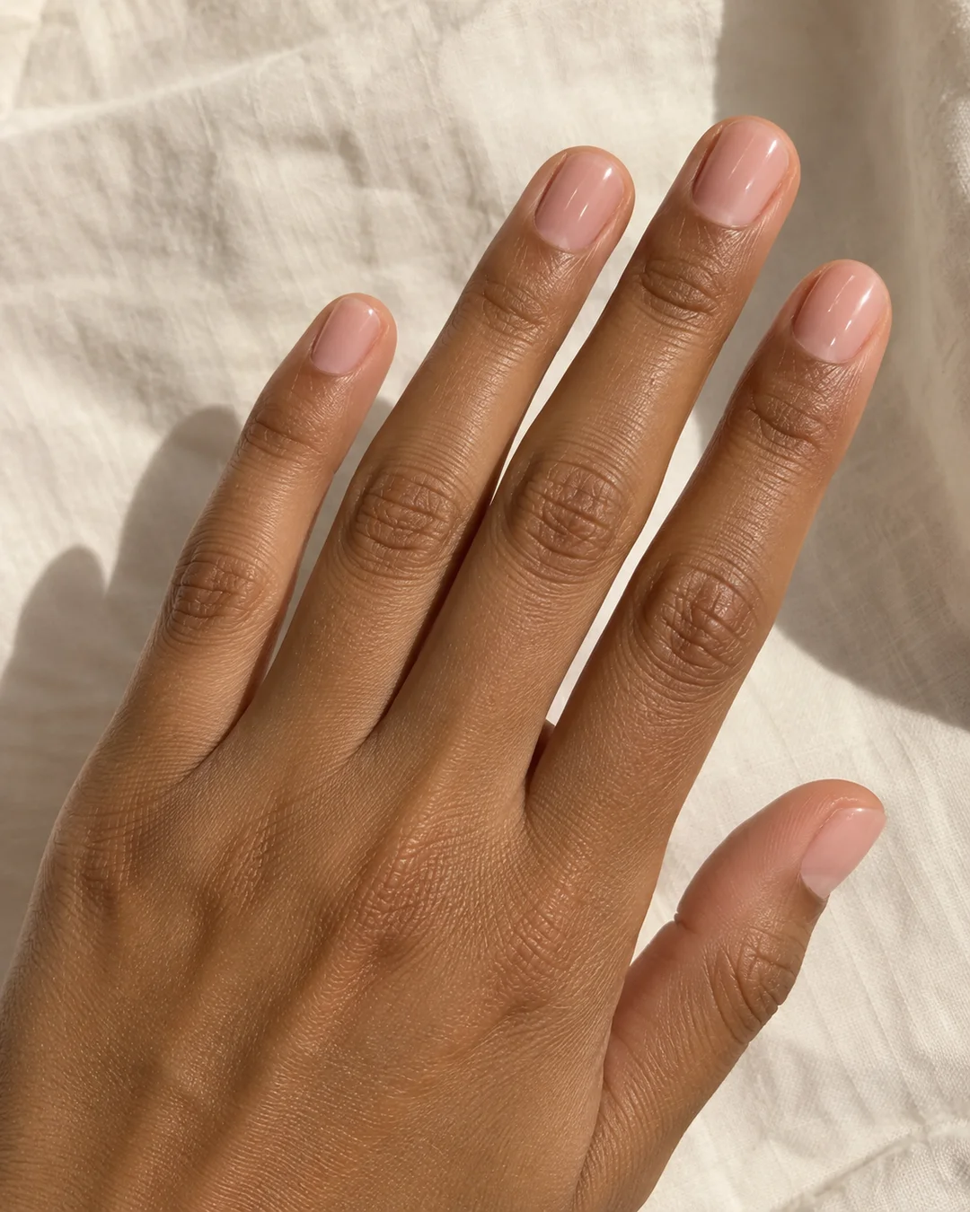 Mano adulta de piel marrón con cinco uñas muy cortas nude rosado, contorno redondeado y brillo liso sobre lino marfil.