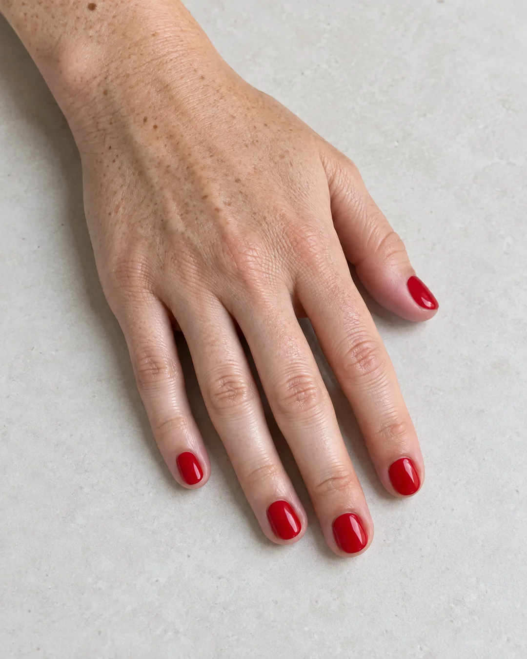 Mano adulta de piel clara con pecas y cinco uñas cortas redondeadas en rojo cereza brillante sobre cerámica clara.