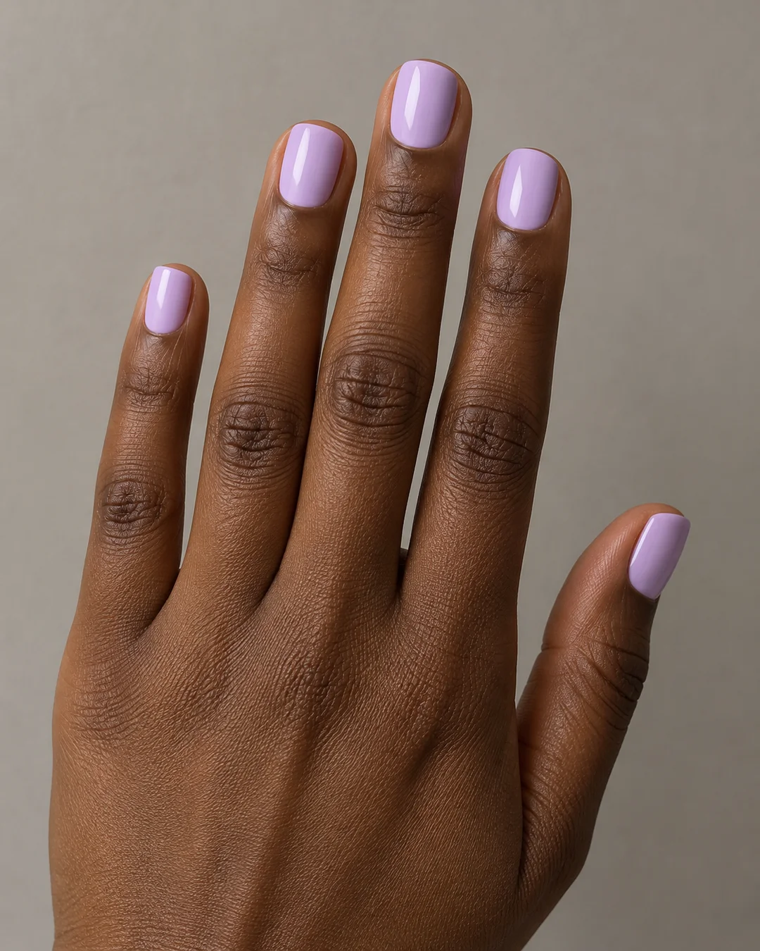 Mano de piel marrón oscura con uñas cortas lila pastel de frente recto y esquinas suaves.