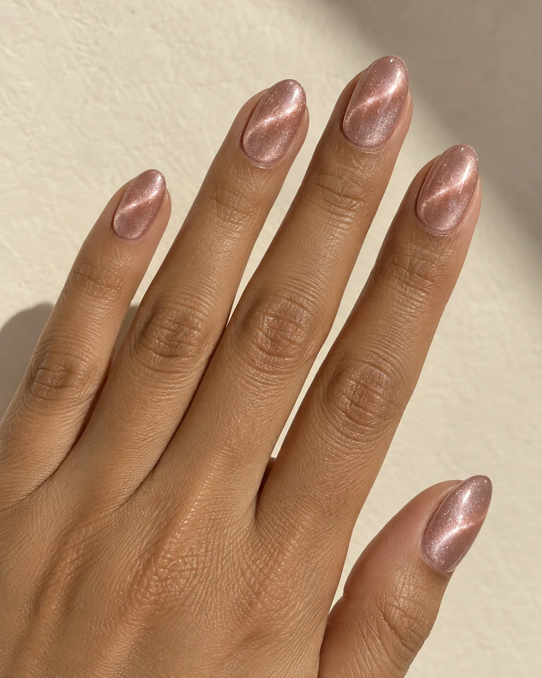Mano adulta de piel oliva con cinco uñas ovaladas medias en rosa champán y bandas luminosas curvas sobre papel crema.