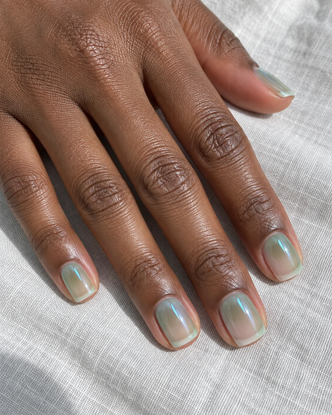 Mano de piel marrón oscura con uñas squoval cortas en verde esmeralda translúcido con reflejo verde azulado y brillo alto.